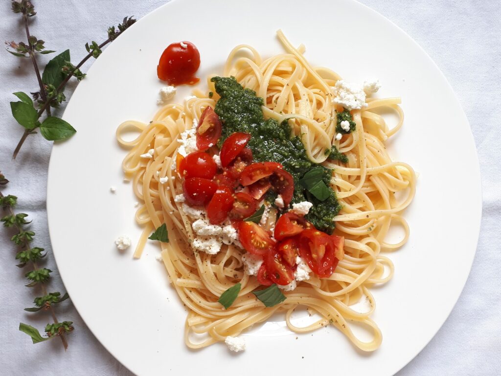 Eine Portion Tagliatelle mit geviertelten Tomaten, Pesto und Schafskäse liegt auf einem weißen Teller.