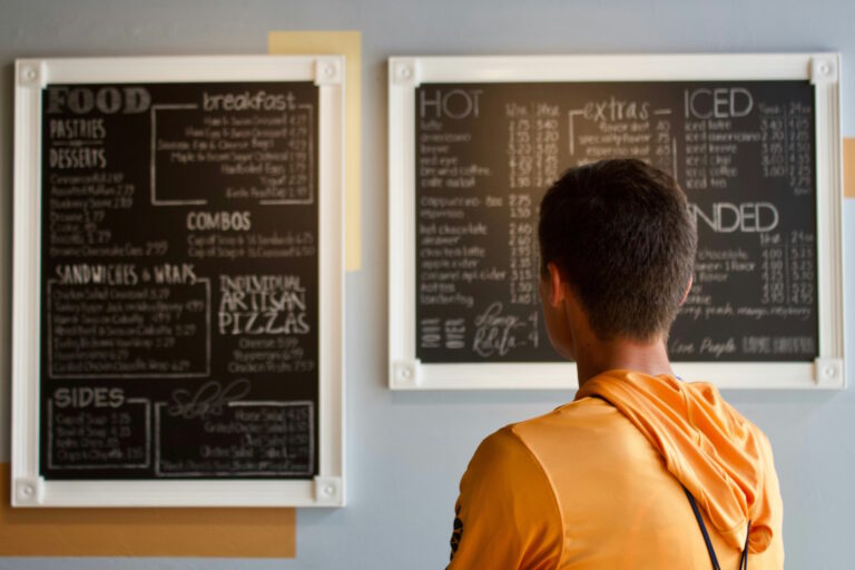 Eine Person betrachtet das Angebot an Speisen und Getränken auf zwei Wandtafeln.