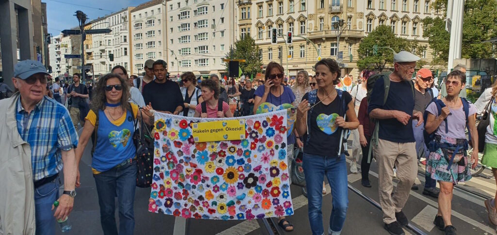 Menschen auf einer Demonstration tragen ein Banner zur Aktion "Häkeln gegen Ökozid"
