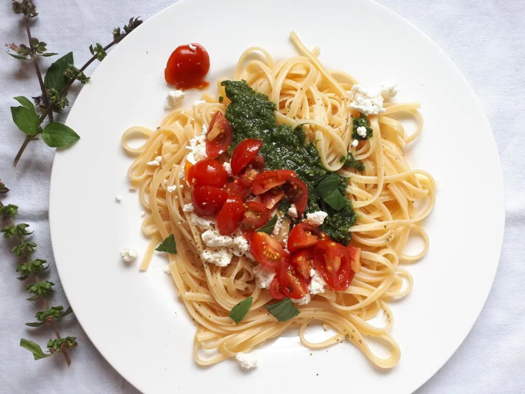 Nudeln mit Pesto und Tomaten Eine Portion Tagliatelle mit geviertelten Tomaten, Pesto und Schafskäse liegt auf einem weißen Teller.