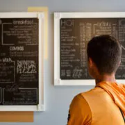 Eine Person betrachtet das Angebot an Speisen und Getränken auf zwei Wandtafeln.